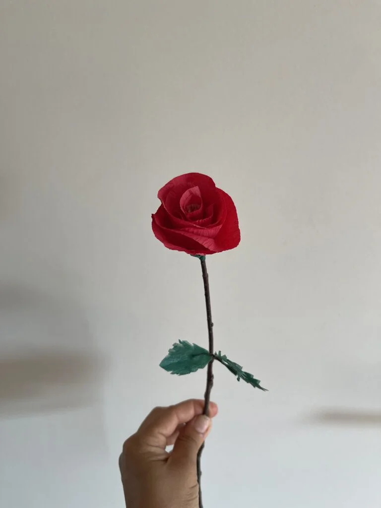 Rosa artesanal terminada de papel crepé con pétalos rojos y hojas verdes sobre una rama natural